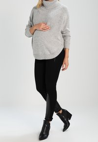 Hellgrauer Strickpullover mit hohem Kragen, geschwunghem Saum und gesprenkelter Textur, kombiniert mit schwarzen Leggings mit Lederakzenten und schwarzen Knöchelstiefeln.