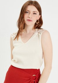 Top sans manches crème avec décolleté en V, présentant des détails en dentelle sur les épaules, associé à une jupe rouge texturée avec un accent de nœud.