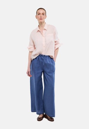 Femme debout les mains dans les poches, portant une chemise rayée rose clair, un pantalon large bleu et des ballerines tressées marron sur fond blanc.