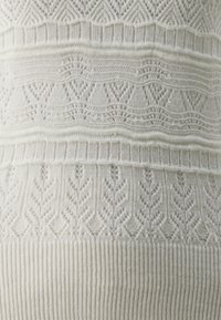 Tissu tricoté couleur crème présentant des motifs floraux et géométriques. Bordure en côtes texturées en bas, mettant en valeur une dentelle complexe.