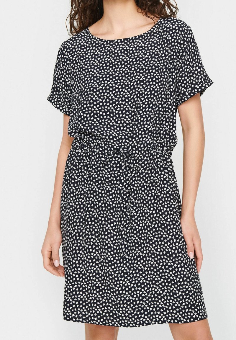 Femme portant une robe noire mi-longue à manches courtes avec un petit motif floral blanc, cintrée à la taille avec un cordon.