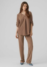 Blusa marrone a maniche lunghe con scollatura a bottoni, abbinata a pantaloni larghi marroni e scarpe con tacco alto blu chiaro. Texture liscia.