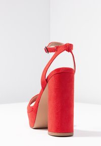 Sandale à talons hauts en daim rouge avec un épais talon bloc et une bride de cheville. L'intérieur est beige et le talon mesure environ 10 cm.