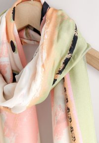 Sciarpa leggera multicolore con fantasie in tonalità pastello pesca, verde e nero, appoggiata con disinvoltura su una gruccia di legno, con una texture morbida.