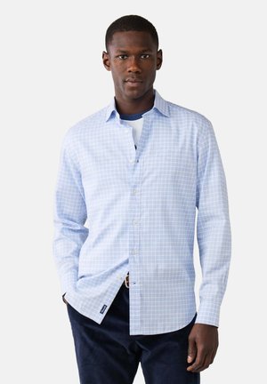Hombre con camisa de cuadros azul claro sobre camiseta blanca y pantalones oscuros, de pie con una mano en el bolsillo frente a fondo blanco.