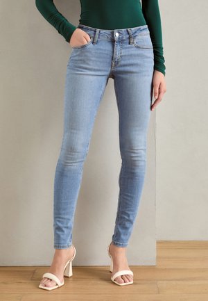Vrouw die lichtblauwe skinny jeans, een groen shirt met lange mouwen en witte sandalen met open teen en blokhak draagt, staand op een houten vloer.