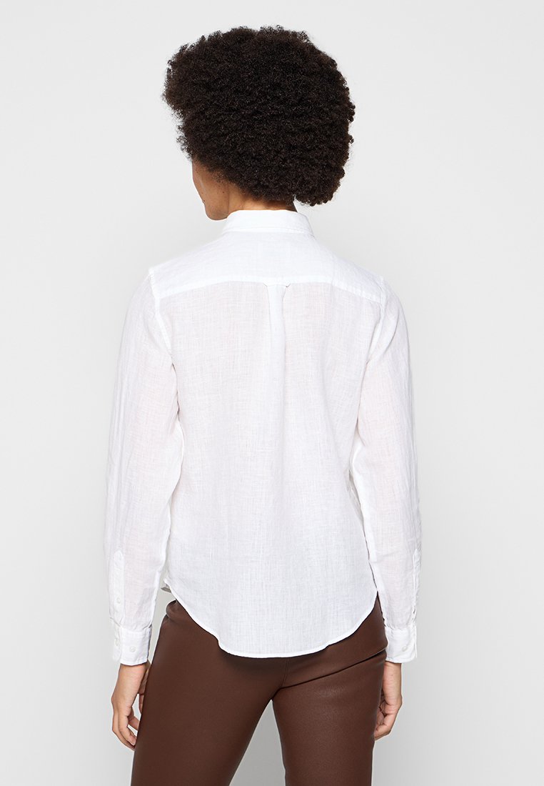 Biała, długa koszula z długim rękawem, wykonana z lekkiej, teksturowanej tkaniny, z zaokrąglonym dołem i zapięciem na guziki przy mankietach, wyeksponowana z tyłu.