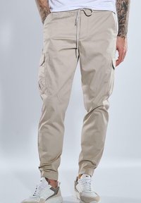 Beige Cargo-Hosen mit elastischem Bund und Kordelzug, ausgestattet mit Seitentaschen und schmal zulaufenden Säumen. Tragen mit weißen und grauen Sneakers.
