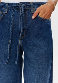 Tummansiniset farkut, joissa on sidottava vyötärö, etutasku ja kontrastivärinen ommel. Kankaalla on sileä pinta.