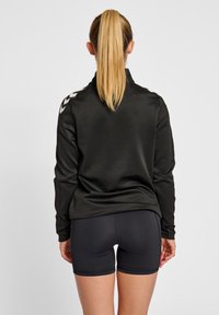 Zwarte sportjack met rits, gladde textuur en wit chevronpatroon op de schouders; gecombineerd met zwarte fitted shorts.