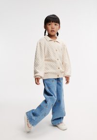 Cardigan crème avec une texture en losanges et de grands boutons, associé à un jean bleu clair avec de petits motifs floraux et des chaussures blanches.