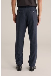 Bărbat purtând pantaloni bleumarin cu nasturi în spate și pantofi negri de costum, stând în fața unui fundal simplu bej.