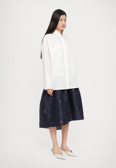 White button-up shirt with two chest pockets, paired with a navy skirt featuring a subtle floral pattern. Flat, pointed beige shoes.