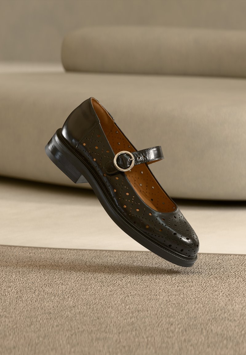 Μαύρο δερμάτινο παπούτσι Mary Jane με διάτρητο σχέδιο, στρογγυλή μύτη, στρογγυλή ασημένια αγκράφα και χαμηλό χοντρό τακούνι, σε ουδέτερο φόντο.