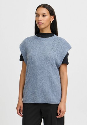 Blue knitted sleeveless sweater with a textured pattern, wide shoulder design, and ribbed neck. Worn over a black top, styled with dark pants.