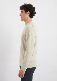 Pull beige à manches longues avec un col rond, poignets côtelés et texture lisse, associé à un pantalon sombre, présenté de côté.