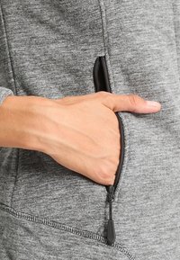 Ręka częściowo wsunięta w zapinaną na zamek boczną kieszeń szarego, strukturalnego, długiego rękawa odzieży.