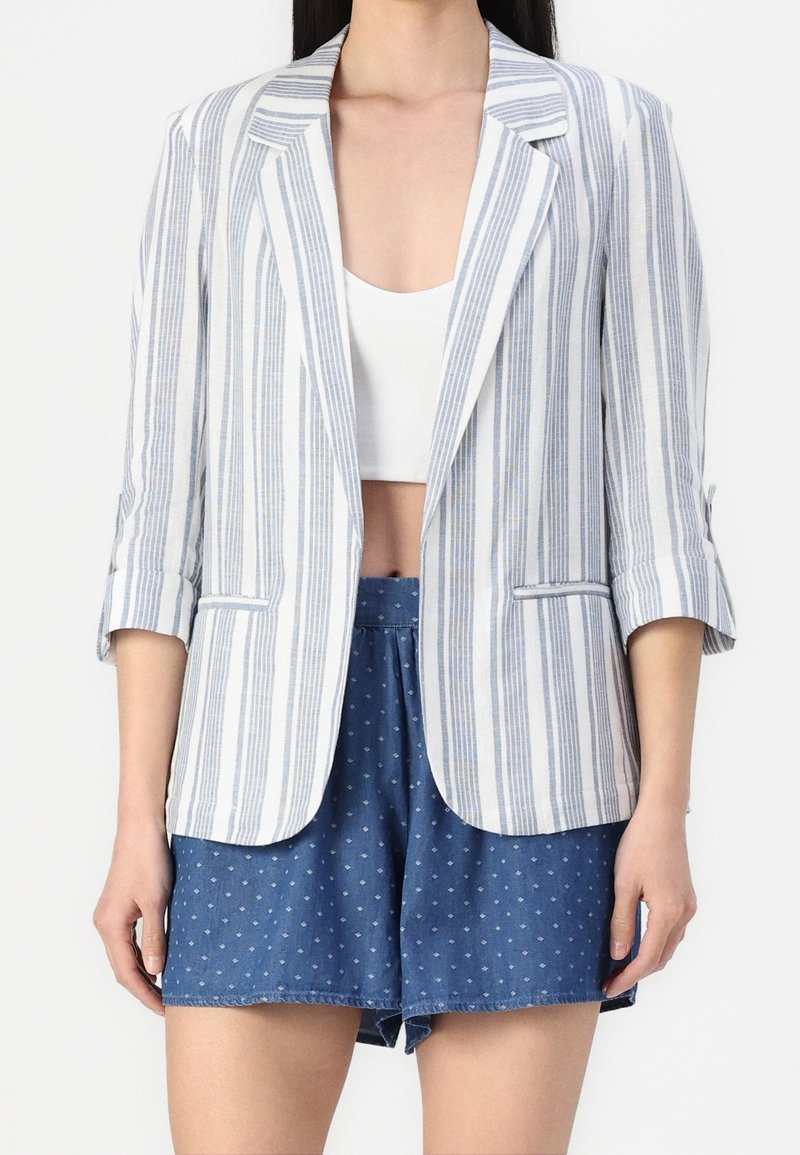 Žena v modro-bíle pruhovaném blejzru přes bílé crop top a modrých vzorovaných šortkách.