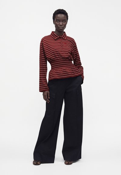 Femme portant un haut à manches longues rayé rouge et blanc avec une taille froncée et un pantalon cargo noir à jambes larges, debout avec une main dans la poche.