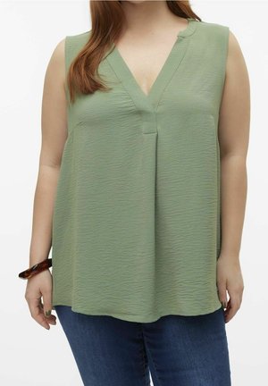 Ärmellose, hellgrüne Bluse mit Textur und V-Ausschnitt, getragen mit dunkelblauen Jeans, ergänzt durch ein Armband aus Schildpatt am linken Handgelenk.