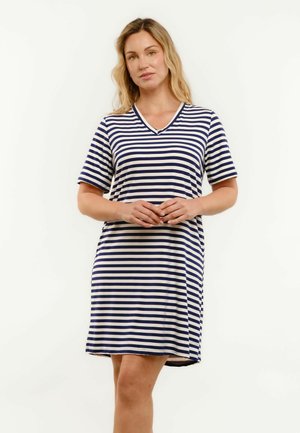 Kobieta o długich blond włosach ubrana w granatowo-białą sukienkę w poziome paski z krótkim rękawem, stojąca z rękami złożonymi z przodu.