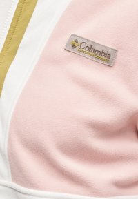 Κοντινή λήψη υφάσματος φλις σε ροζ χρώμα με λο γότυπο της εταιρείας Columbia Sportswear και δίπλα φερμουάρ λευκό με κίτρινη ραφή.