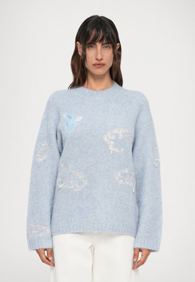 Femeie îmbrăcată cu un pulover bleu deschis, cu nori albi brodați și o pasăre albastră, asortat cu pantaloni albi, stând în fața unui fundal simplu.