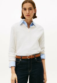 Pull en V texturé blanc avec des manches longues, superposé sur une chemise à col bleu clair. Porté avec un jean foncé et une ceinture marron.