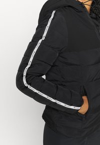 Svart puffjacka med huva, med en elegant design, texturerat tyg och kontrasterande vitt logoband längs ärmarna.