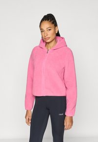 Bluza z polaru z kapturem na zamek w jasnoróżowym kolorze o przytulnej fakturze i kapturze. Posiada pełny przedni zamek oraz ściągacze na mankietach. Noszona z czarnymi legginsami.