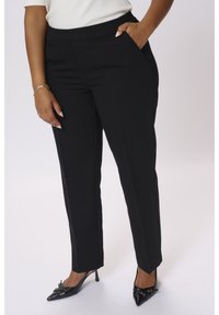 Femme portant un pantalon noir droit avec des poches latérales, associé à des talons noirs pointus et un haut blanc.