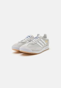 Szare zamszowe sneakersy z białymi skórzanymi akcentami, trzema paskami i niebieskimi sznurówkami. Posiadają białą gumową osłonę na palce oraz teksturowaną gumową podeszwę.