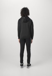 Μαύρο hoodie με ένα αντίστοιχο σετ φόρμας. Το sweatshirt διαθέτει κουκούλα και οι φόρμες έχουν λευκές πλευρικές ρίγες. Το υλικό φαίνεται λείο και ελαστικό.