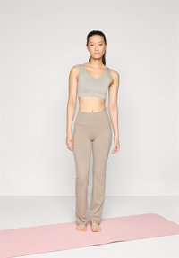 Top corto gris claro y leggings beige hechos de una tela lisa y elástica. La modelo está de pie sobre una esterilla de yoga rosa, descalza, con un fondo neutro.