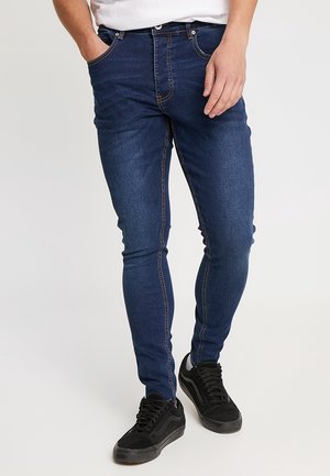 Man draagt donkerblauwe skinny jeans en zwarte sneakers, staand met één hand in de zak, tegen een effen lichte achtergrond.