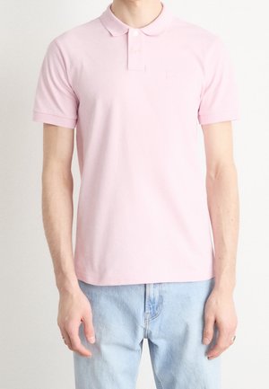 Hellrosa Poloshirt mit klassischem Kragen, kurzen Ärmeln und einem dezenten Logo auf der Brust, kombiniert mit hellblauer Jeans.