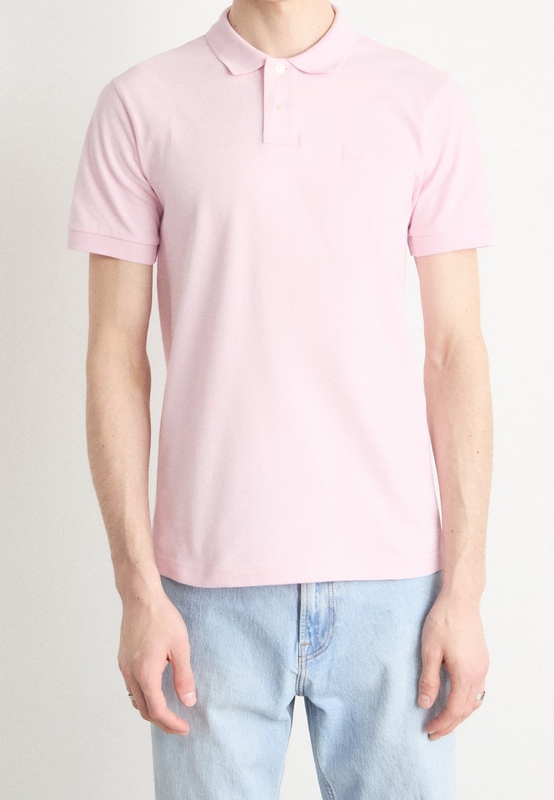 Polo rose clair avec col classique, manches courtes et un logo discret sur la poitrine, associé à un jean en denim bleu clair.