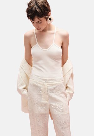 Donna con capelli corti indossa una canottiera bianca, pantaloni bianchi strutturati, una giacca abbinata drappeggiata sulle braccia e un grande orecchino a forma di fiore bianco.