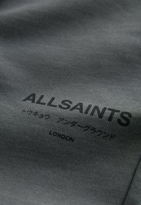 Sivkasta tkanina z gladko teksturo prikazuje črn tisk: "ALLSAINTS," japonska fraza in "LONDON" natisnjeno spodaj.