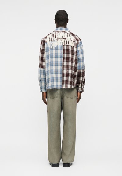 Άνδρας που φοράει πουκάμισο μπλε και καφέ καρό με το κείμενο "Billionaire Boys Club" στην πλάτη, χαλαρό γκρι παντελόνι και μαύρα παπούτσια, με την πλάτη προς το μέρος μας.