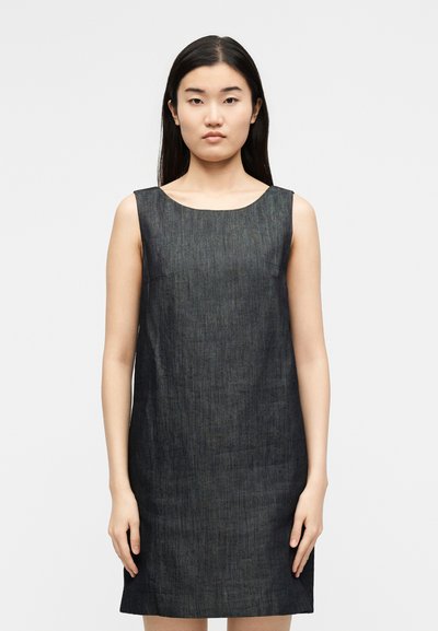 Γυναίκα με μακριά σκούρα μαλλιά που φορά ένα αμάνικο, σκούρο μπλε τζιν φόρεμα, στέκεται μπροστά σε ένα απλό ανοιχτό γκρι φόντο.