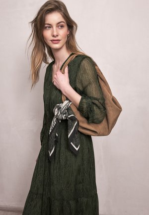 Jeune femme aux cheveux clairs portant une robe verte à motifs, tenant un sac en daim beige sur lequel est noué un foulard noir et blanc.