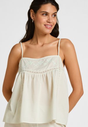 Vrouw draagt een mouwloze, crèmekleurige top met dunne bandjes, bloemrijke borduursels en kantdetails, glimlacht en kijkt naar de zijkant.