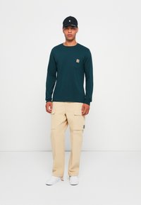 Donkergroene longsleeve shirt met een voorzak en beige cargobroek met zijzakken, gecombineerd met witte schoenen en een zwarte pet.