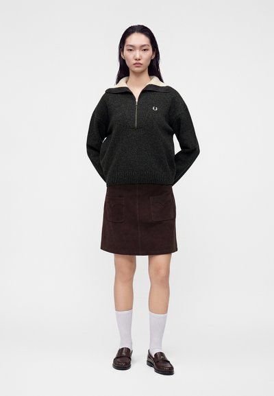 Jeune femme debout portant un pull sombre à demi-fermeture éclair, une jupe marron avec poches avant, des chaussettes blanches et des mocassins foncés sur un fond uni.