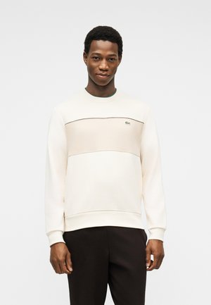 Νέος άνδρας φορώντας ένα εκρού φούτερ με μπεζ πάνελ και μικρό λογότυπο, συνδυασμένο με μαύρο παντελόνι, στέκεται μπροστά σε απλό λευκό φόντο.