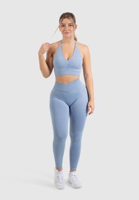 Lichtblauwe sportbeha met V-hals en geplooide details, gecombineerd met high-waisted leggings in dezelfde tint, gedragen met witte sportschoenen.