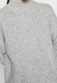 Grå stickad tröja med hög krage, med ett texturerat mönster och lätt ribbning vid axlarna. Mjuk, mysig tyg.