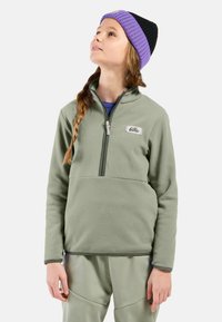Pull en polaire vert clair avec un col zippé mi-hauteur, une poche avant et des accents vert olive. Porté avec un bonnet violet et noir.