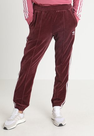 Person trägt burgunderrote Adidas-Trainingshose mit weißen Streifen, elastischen Bündchen und weißen Adidas-Sneakern, steht mit den Händen in den Taschen.